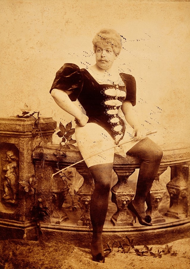 Man seated, wearing corset and holding whip